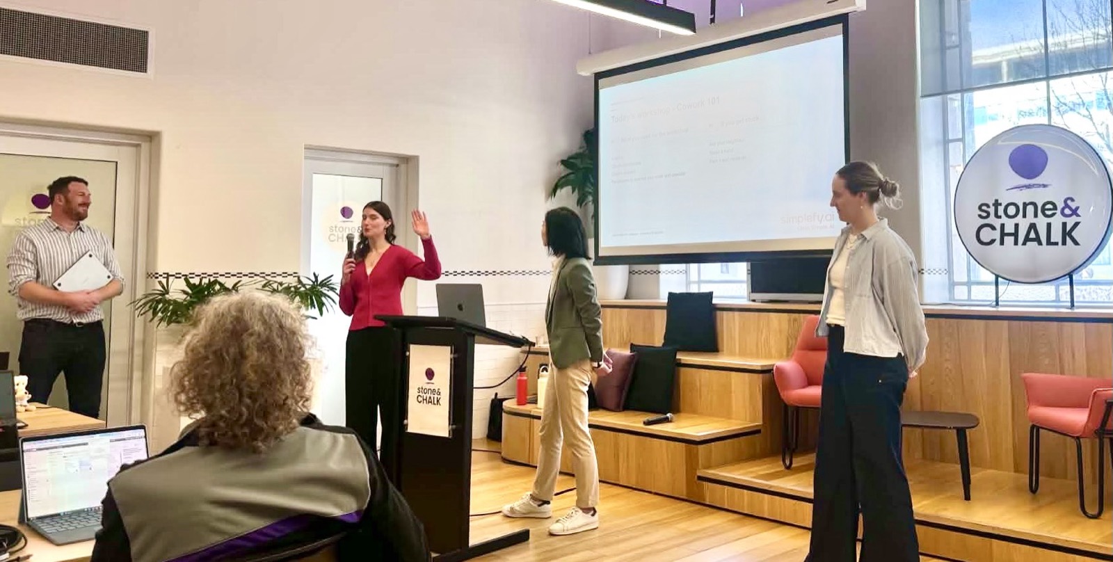Jess Tresidder presenting from a lectern at Stone and Chalk with two facilitators helping the room.
