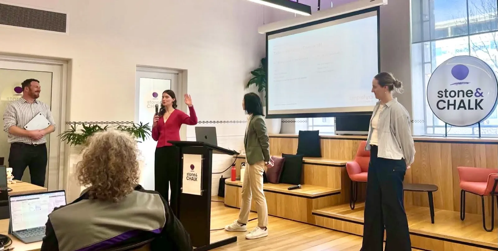 Jess Tresidder presenting from a lectern at Stone and Chalk with two facilitators helping the room.
