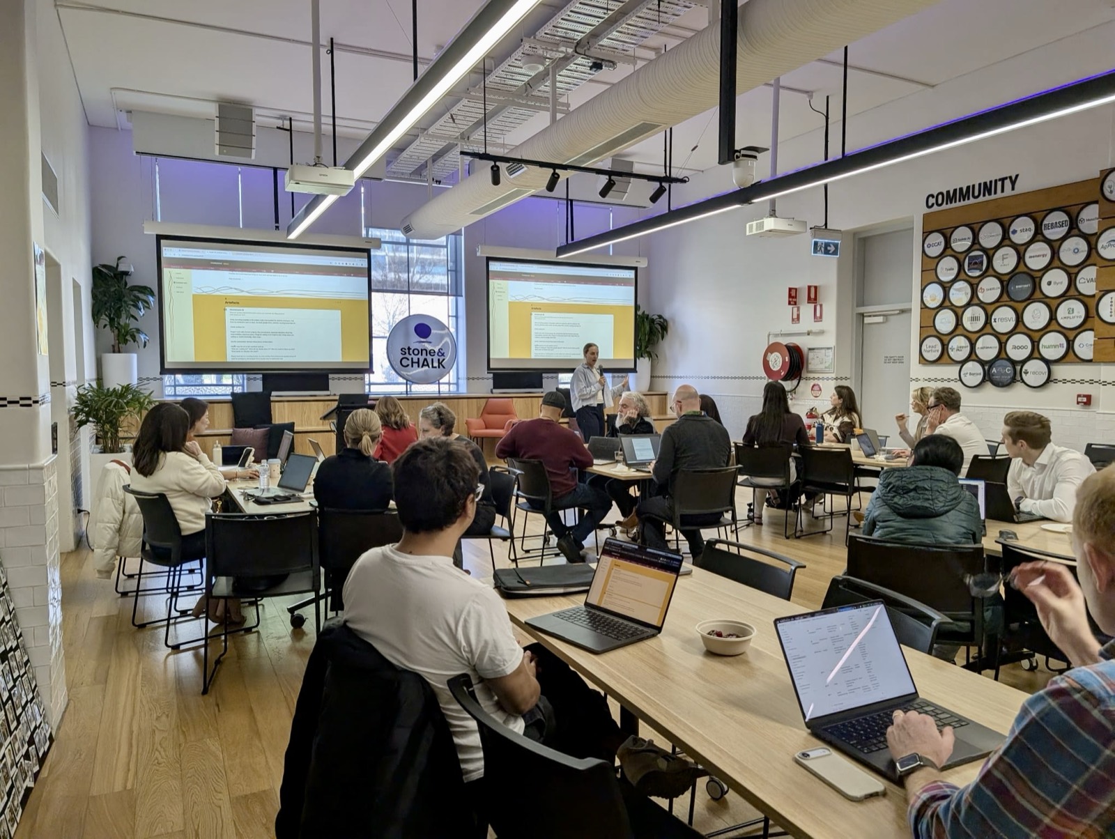 A full room of attendees with laptops open at the Stone and Chalk Claude 101 workshop, two screens showing the session slides.