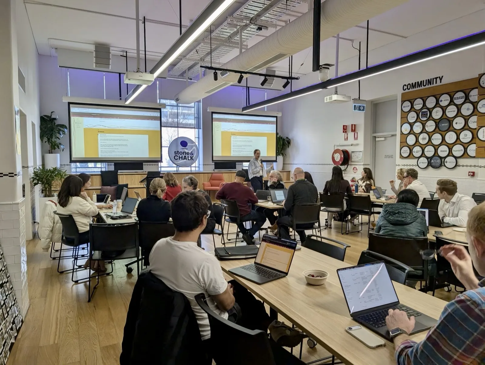 A full room of attendees with laptops open at the Stone and Chalk Claude 101 workshop, two screens showing the session slides.